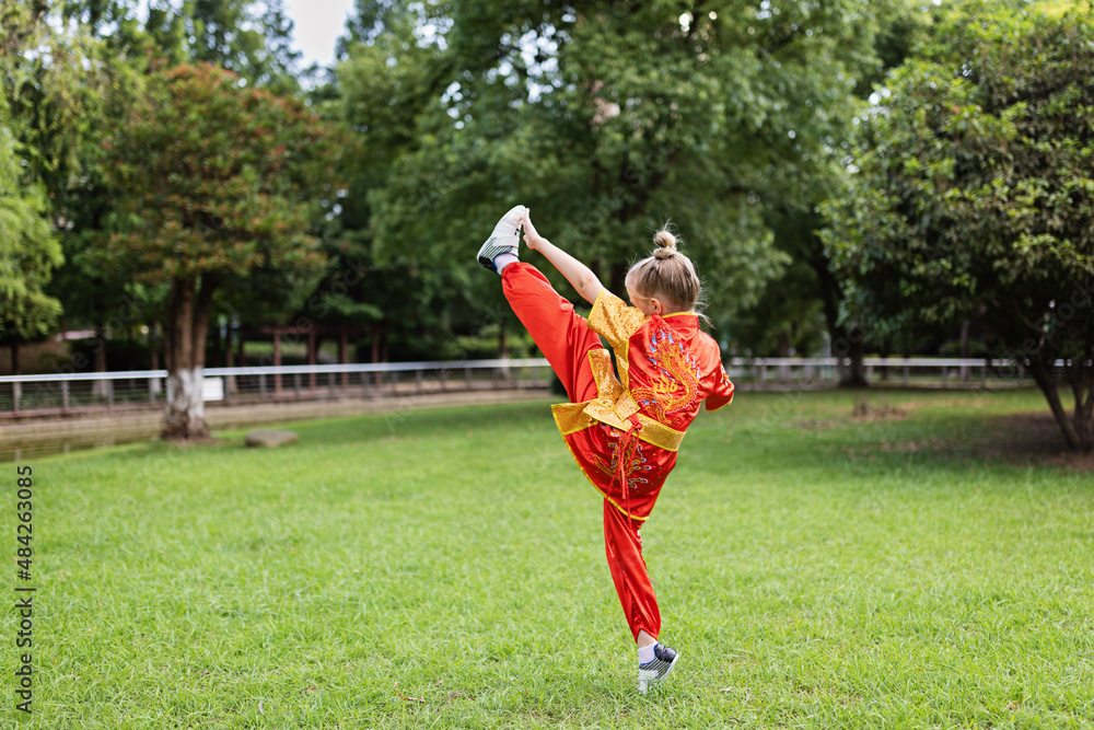 Obraz premium Cute little caucasian girl seven years old in red sport wushu uniform exercising in park at summer day. Lifestyle portrait of kung fu fighter child athlete