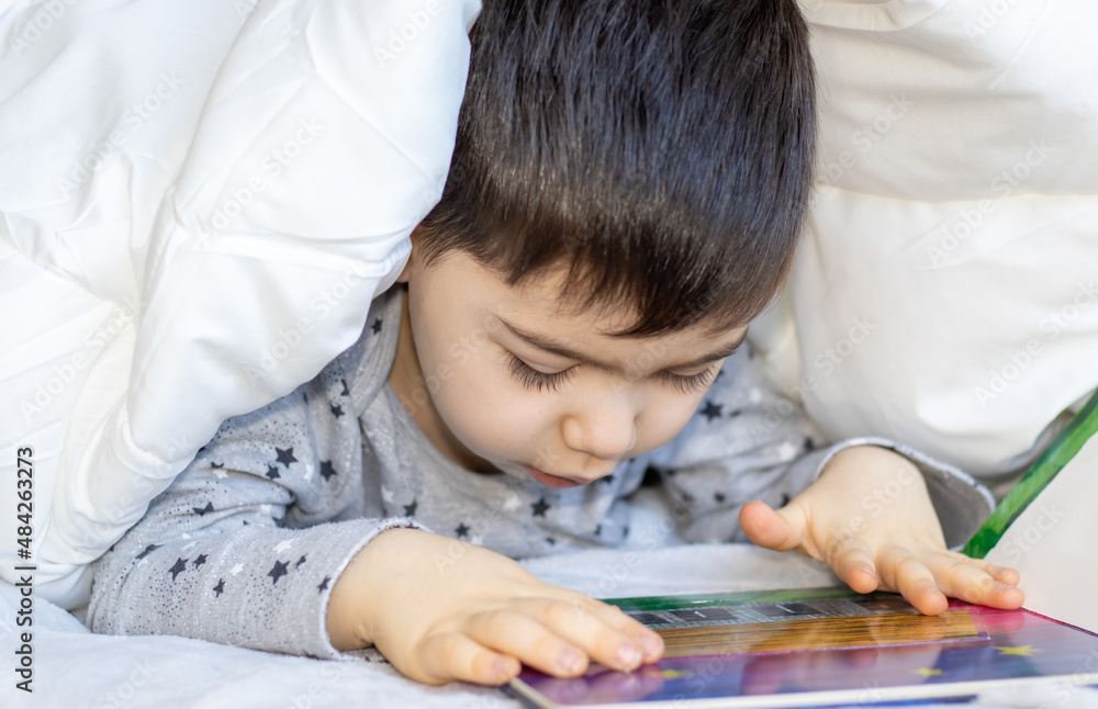 cute kid covered with white soft blanket is reading a book for kids. little boy is looking at the camera or in the book. cozy atmosphere. activities for kids. reading books. cute kid covered with white soft blanket is reading a book for kids. little boy is looking at the camera or in the book. cozy atmosphere. activities for kids. reading books.
