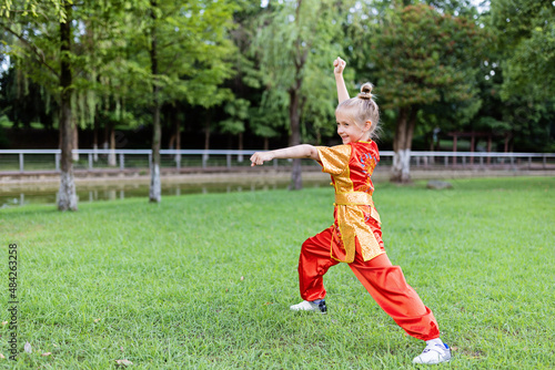 Cute little caucasian girl seven years old in red sport wushu uniform exercising in park at summer day. Lifestyle portrait of kung fu fighter child athlete