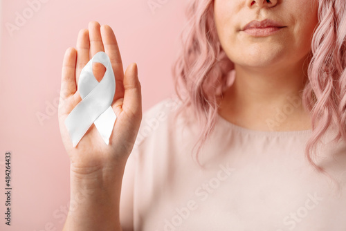 Mental health awareness campaign. Woman hand holding white ribbon on isolated pastel pink background for supporting people living and illness.