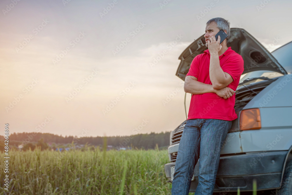 Man talking by phone leaning to van standing on rural road with raised hood. Service and assistance on road. Man talking by phone leaning to van standing on rural road with raised hood. Service and assistance on road.