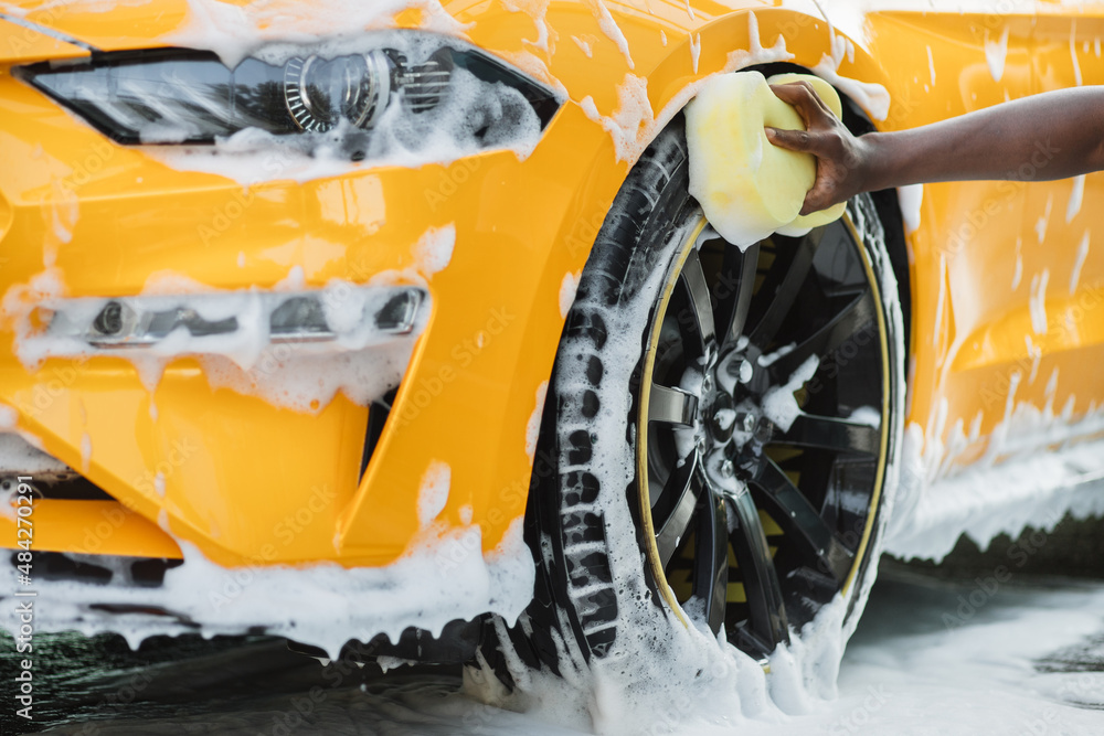 Hands of African man holding yellow sponge, washing car wheel with foam