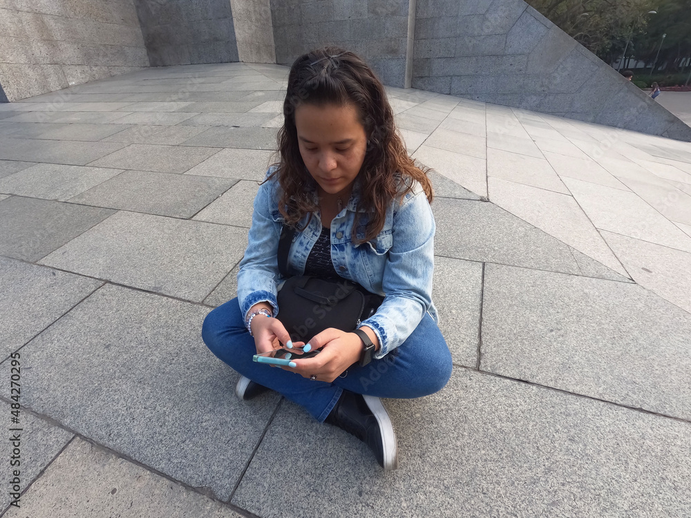 joven mujer sentada en piso con piernas cruzadas revisa su teléfono celular joven mujer sentada en piso con piernas cruzadas revisa su teléfono celular