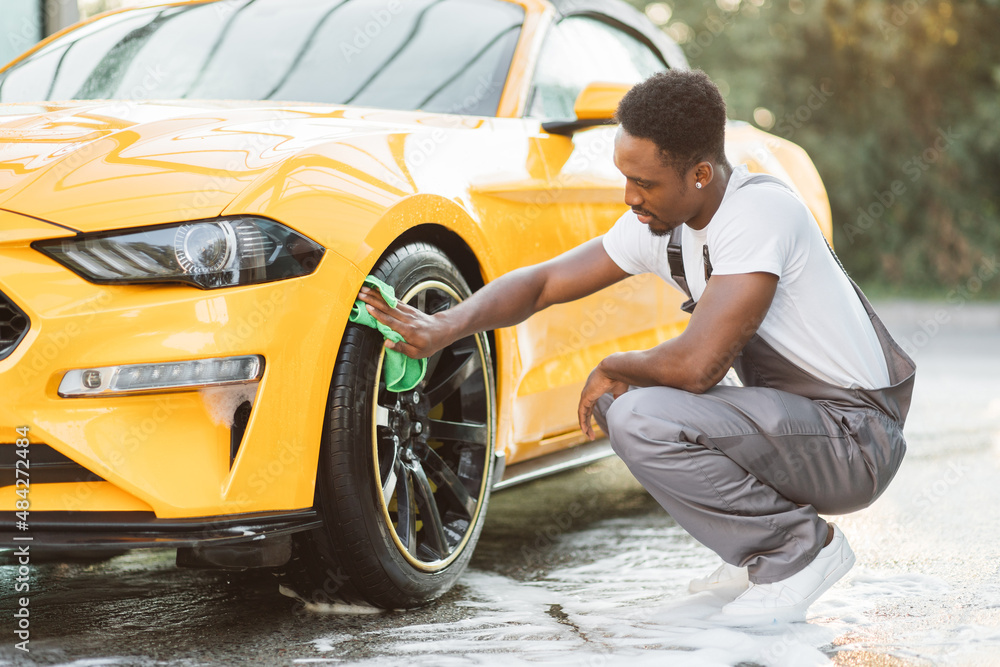 Car wash service outdoors. Car wash self-service concept. Handsome African guy wiping wheel of modern luxury car with green microfiber cloth during the washing process outdoors. Car wash service outdoors. Car wash self-service concept. Handsome African guy wiping wheel of modern luxury car with green microfiber cloth during the washing process outdoors.