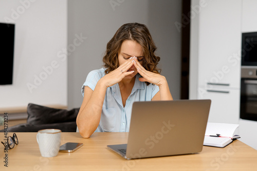 Tired female student feeling bored and tired. Attractive brunette woman sits with eyes closed in front of the laptop, leans head on the hand, feels headache and burnout