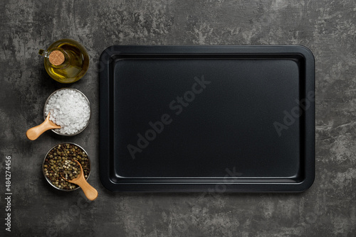 Empty baking sheet, olive oil jar, salt and pepper grains over black textured background. Rectangular oven tray and spices for baking and roasting. Baking pan for cooking and food design. photo