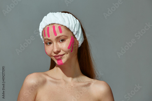 Beautiful redhead young girl with bare shoulders, face lift tapes on face enjoy facelifting rejuvenating daily procedure