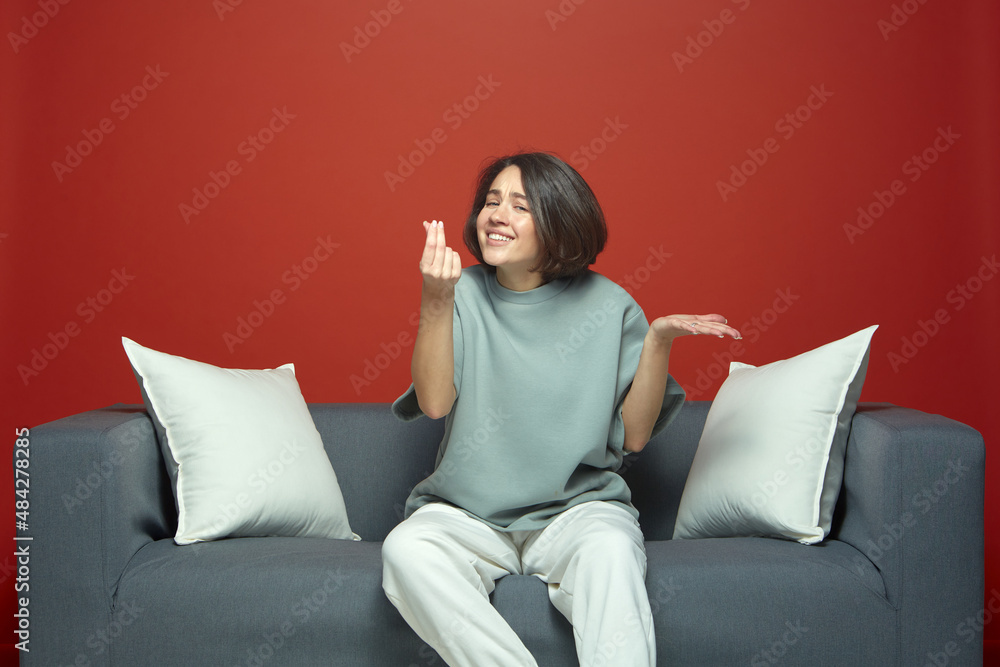 Emotional young beautiful girl show money gesture, asking cash, sitting on sofa. Not enough money. Debt or credit. Emotional young beautiful girl show money gesture, asking cash, sitting on sofa. Not enough money. Debt or credit.