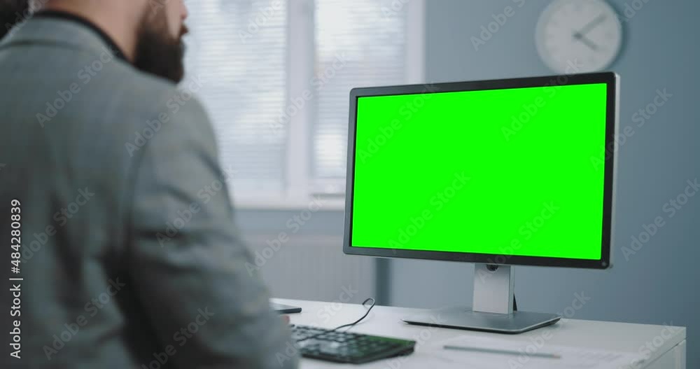 View from shoulder of mature businessman using modern pc with green mock up screen while working at bright office. Focus on chrome key monitor. View from shoulder of mature businessman using modern pc with green mock up screen while working at bright office. Focus on chrome key monitor.