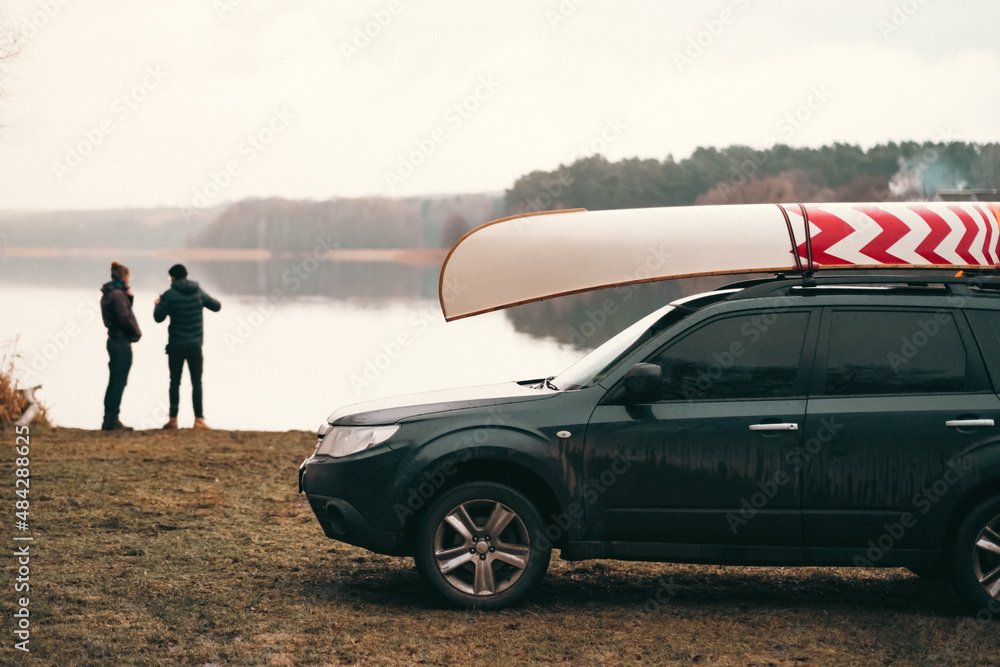 A car with mounted canoe on the rooftop by the lake, two men standing in the background. Autumn or spring nature scene, paddling lifestyle