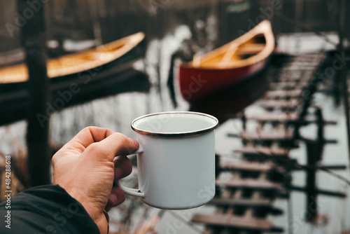 A tin hiking cup in hand with canoe boats in the background  point of view shot. Paddling lifestyle in springtime or autumn season