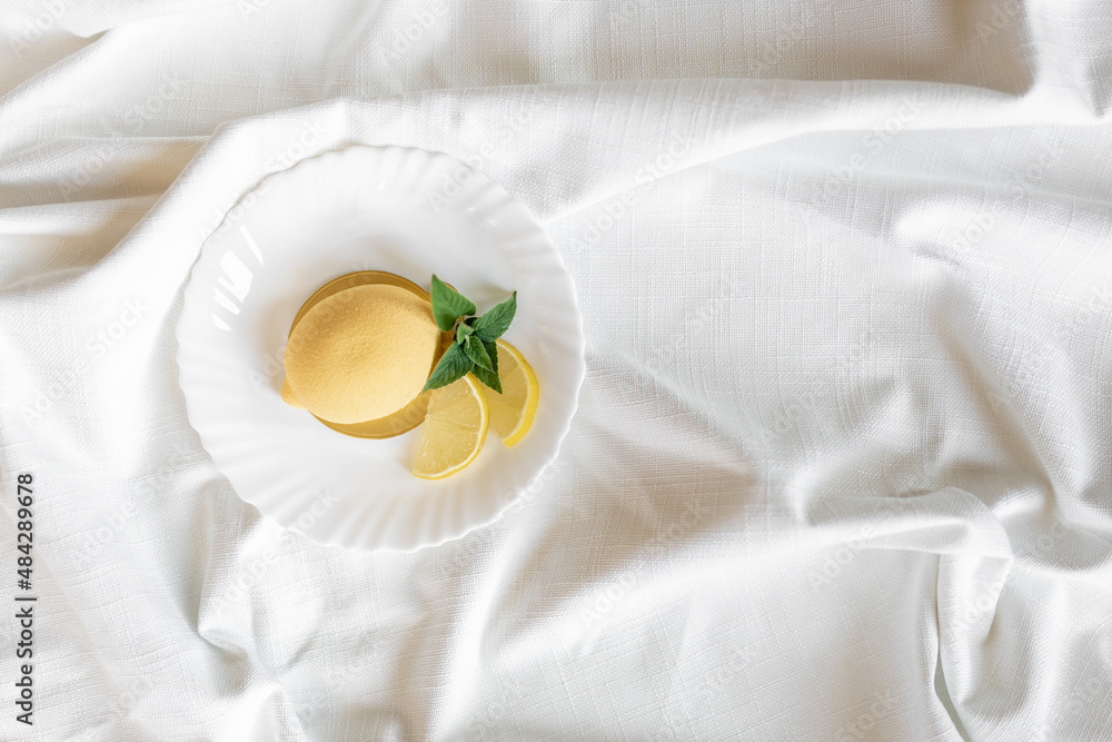 Mousse yellow cake in the form of a lemon with a green mint leaf and real lemon slices on a white plate on a light background. Delicious and beautiful pastries. Background for confectionery Mousse yellow cake in the form of a lemon with a green mint leaf and real lemon slices on a white plate on a light background. Delicious and beautiful pastries. Background for confectionery