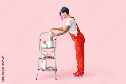 Female painter with ladder and cans on pink background