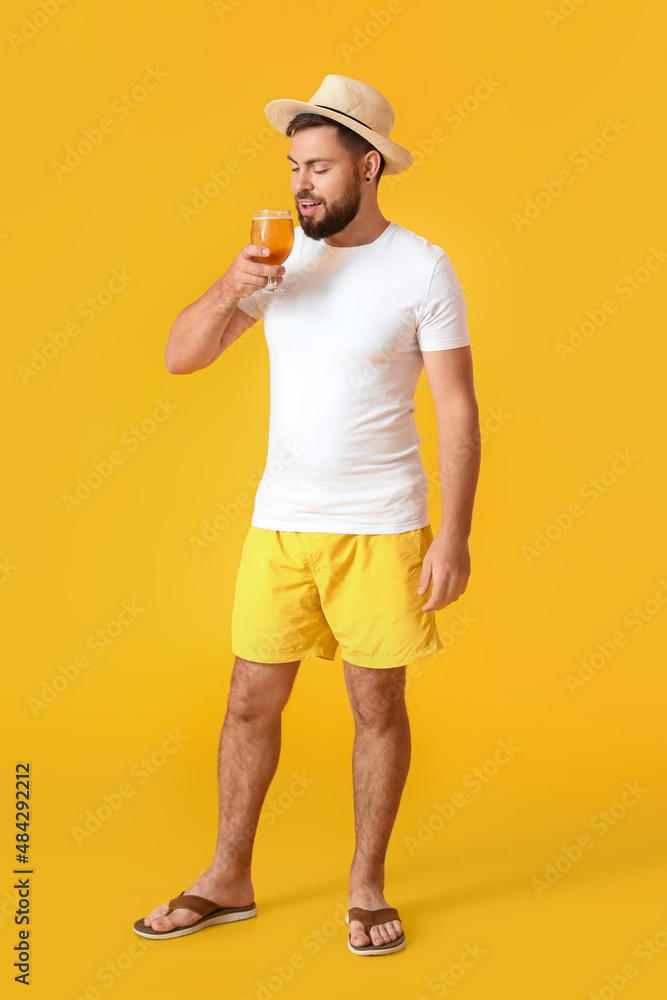Young man in hat drinking beer on yellow background