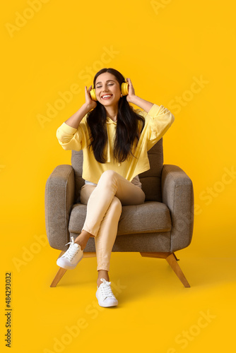 Beautiful young woman with headphones listening to music in soft armchair on yellow background © Pixel-Shot