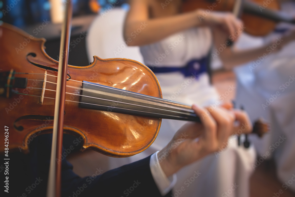 Male violin player, fiddler violinist with a bow performing music on stage during concert with orchestra and musical band in the background Male violin player, fiddler violinist with a bow performing music on stage during concert with orchestra and musical band in the background
