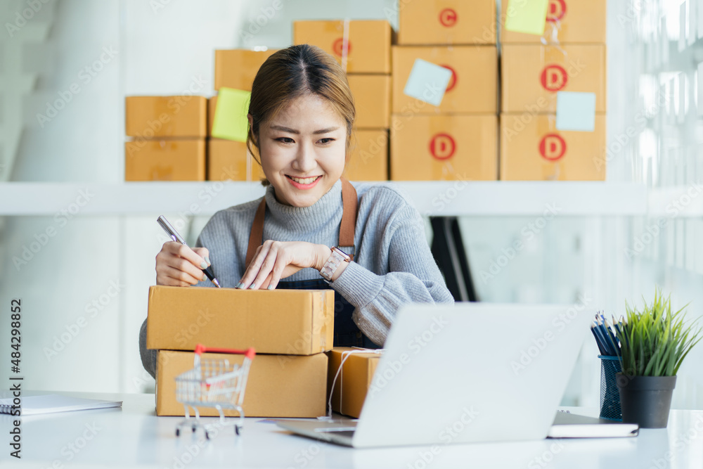 Cheerful female entrepreneur smiling and writing address on carton box while sending order to customer at home Cheerful female entrepreneur smiling and writing address on carton box while sending order to customer at home