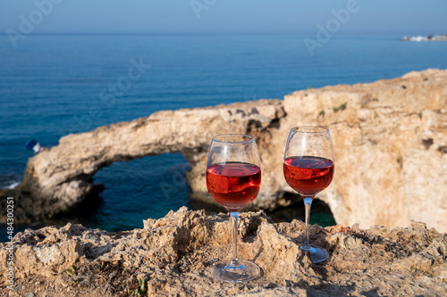 Two glasses of rose dry white wine served on rocks in blue sea bay with Love Bridge on background near Ayia Napa touristic town on Cyprus