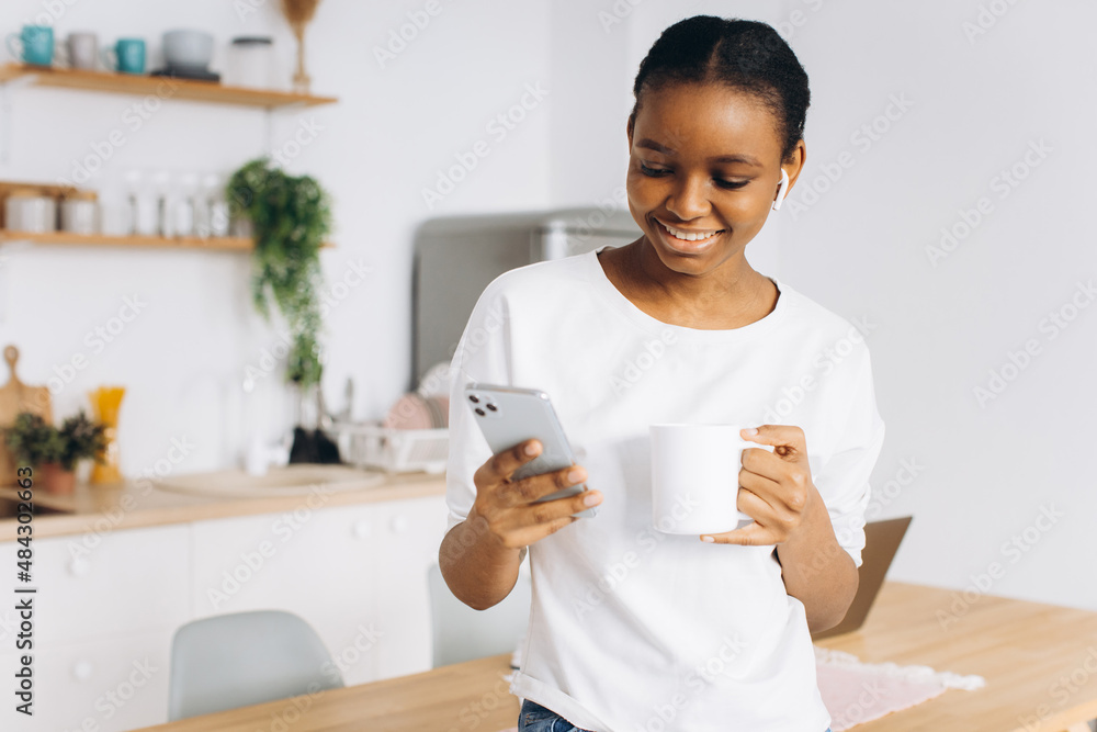 Portrait of a cheerful young black woman holding a phone and drinking coffee in the kitchen. Portrait of a cheerful young black woman holding a phone and drinking coffee in the kitchen.
