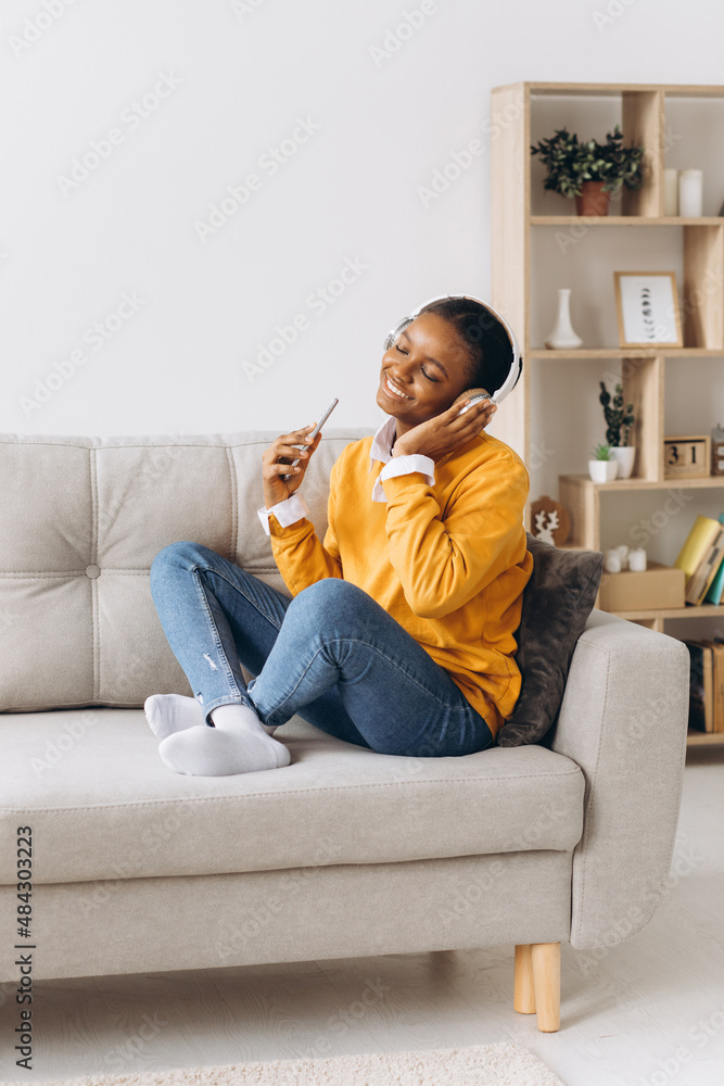 People, technology and leisure concept - happy african american young woman sitting on sofa with smartphone and headphones listening to music at home People, technology and leisure concept - happy african american young woman sitting on sofa with smartphone and headphones listening to music at home