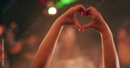 Good music does the heart good. Hands of woman making a heart shape love gesture during a live music performance. Audience having fun and enjoying a festival or party vibe with strobe lights