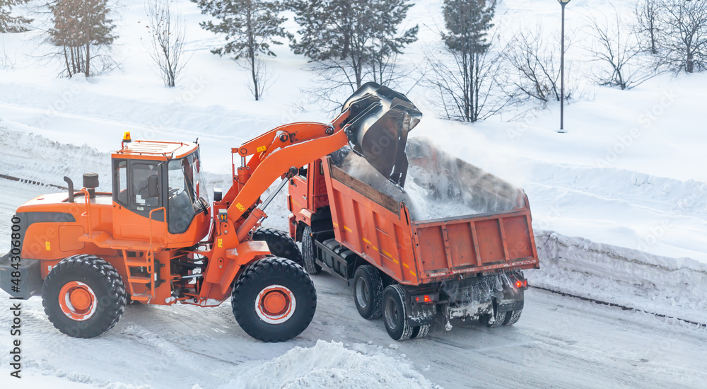 Big orange tractor cleans up snow from the road and loads it into the truck. Cleaning and cleaning of roads in the city from snow in winter. Snow removal after snowfall and blizzards. Big orange tractor cleans up snow from the road and loads it into the truck. Cleaning and cleaning of roads in the city from snow in winter. Snow removal after snowfall and blizzards.