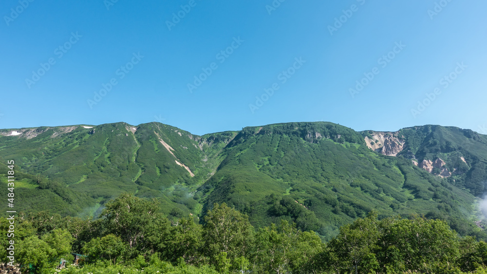 A picturesque mountain range against a clear blue sky. There is lush green vegetation on the slopes. The forest is at the foot. Copy space. Kamchatka