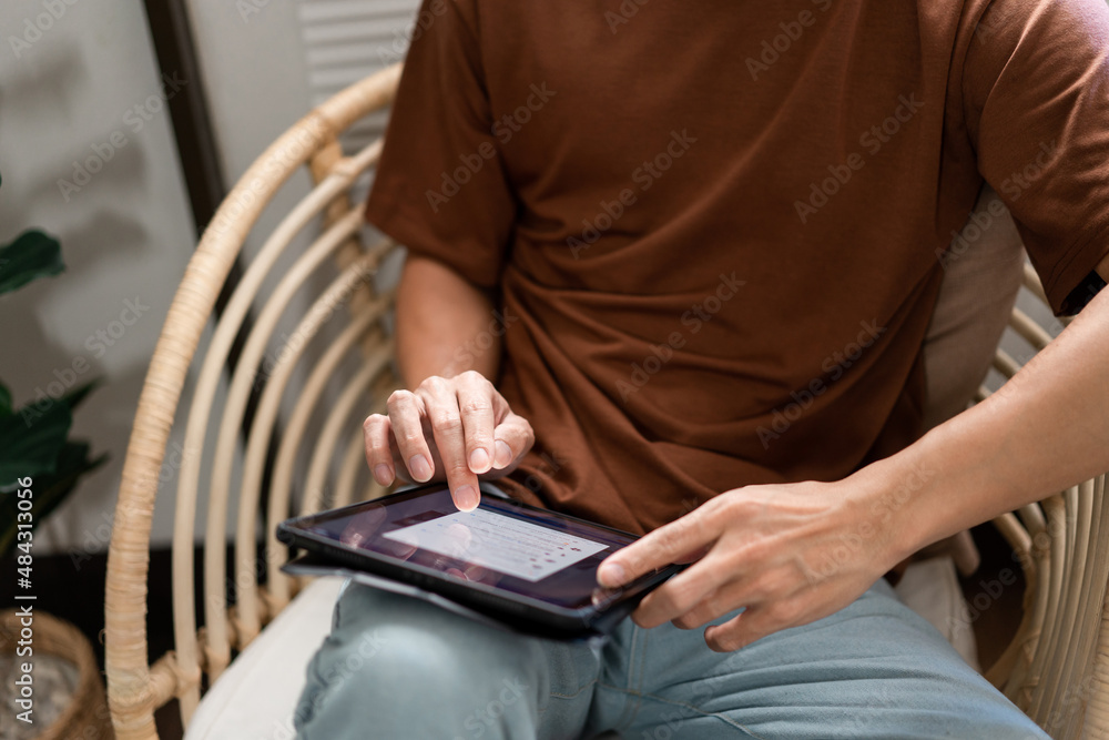 Technology Concept The male with his casual T-shirt and jeans sitting comfortably on the wooden chair and doing touchscreen for checking the web browser on the iPad