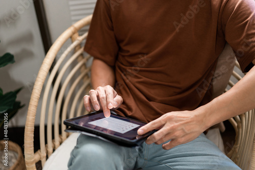 Technology Concept The male with his casual T-shirt and jeans sitting comfortably on the wooden chair and doing touchscreen for checking the web browser on the iPad