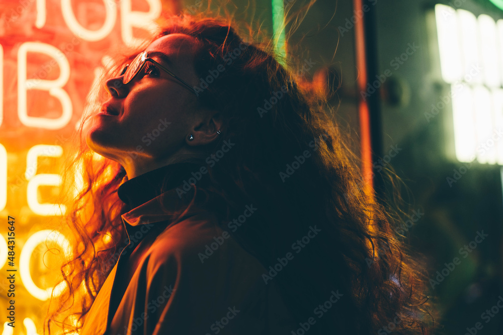 Curly haired young woman tourist with light makeup in glasses looks around standing near bar with colorful neon sign against night megalopolis - obrazy, fototapety, plakaty 