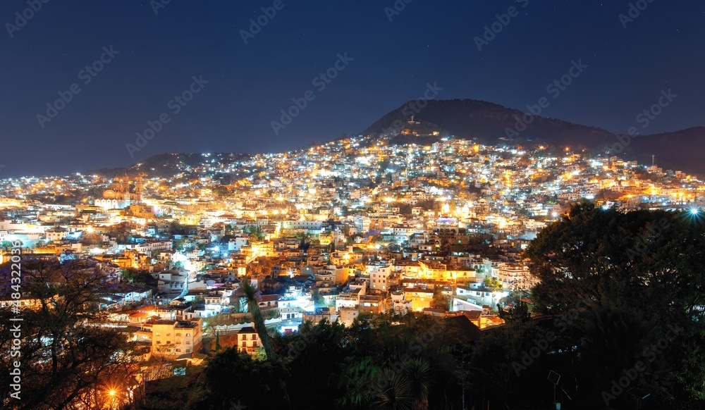 Naklejka premium View of the night city on the slopes of the mountains. Colonial architecture, lots of lanterns and light in the windows. Dark night sky. The cramped streets of Taxco in Mexico