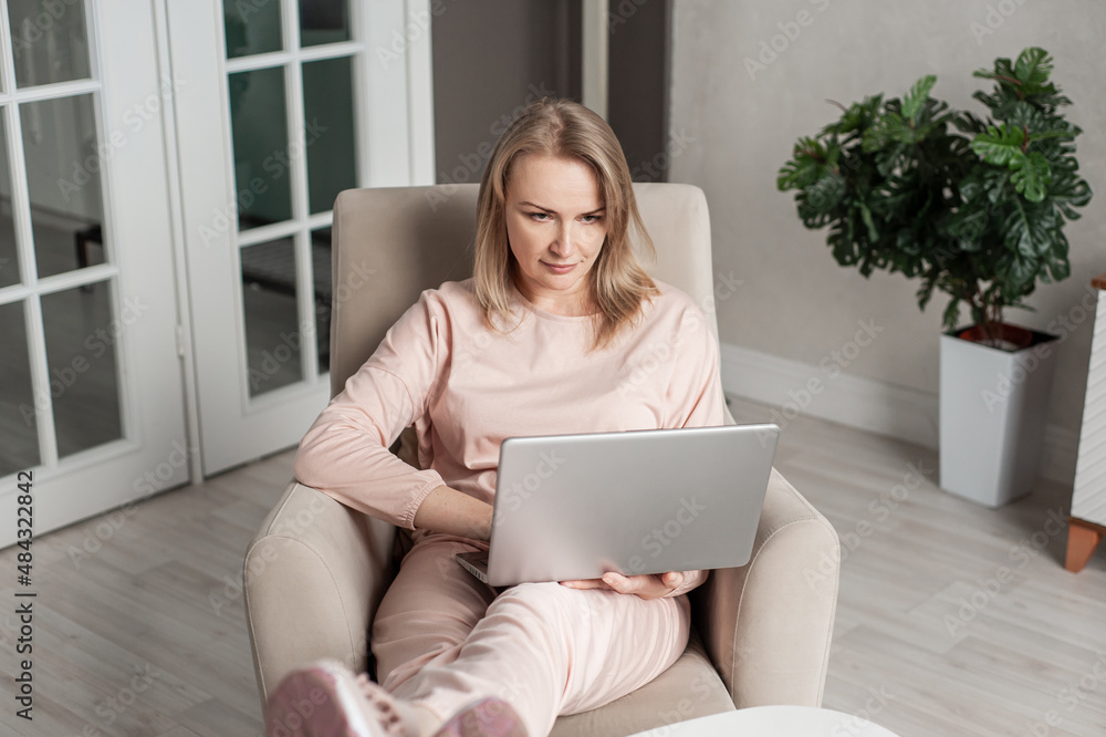 A beautiful blonde middle-aged woman sees in a cozy home chair with a laptop in her hands. Work or study from home. Freelancing or distance learning. A beautiful blonde middle-aged woman sees in a cozy home chair with a laptop in her hands. Work or study from home. Freelancing or distance learning.