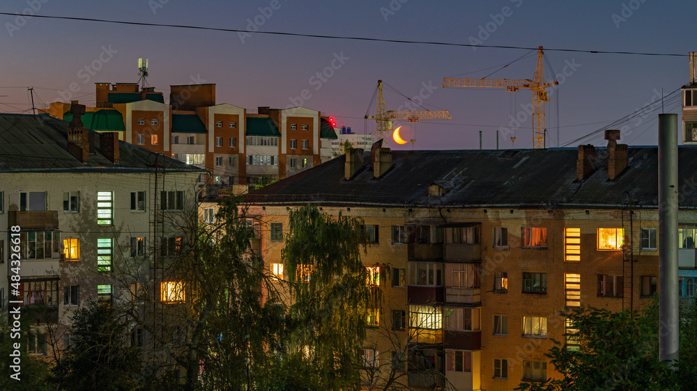 Light in the windows of high-rise buildings at night in summer and half-moon in the sky between houses