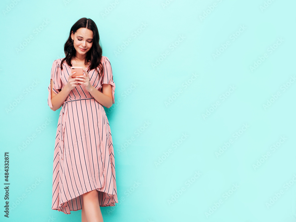 Young beautiful smiling female in trendy summer dress. Sexy carefree woman posing near blue wall in studio. Positive brunette model looking at cellphone screen. Holding smartphone and using apps Young beautiful smiling female in trendy summer dress. Sexy carefree woman posing near blue wall in studio. Positive brunette model looking at cellphone screen. Holding smartphone and using apps