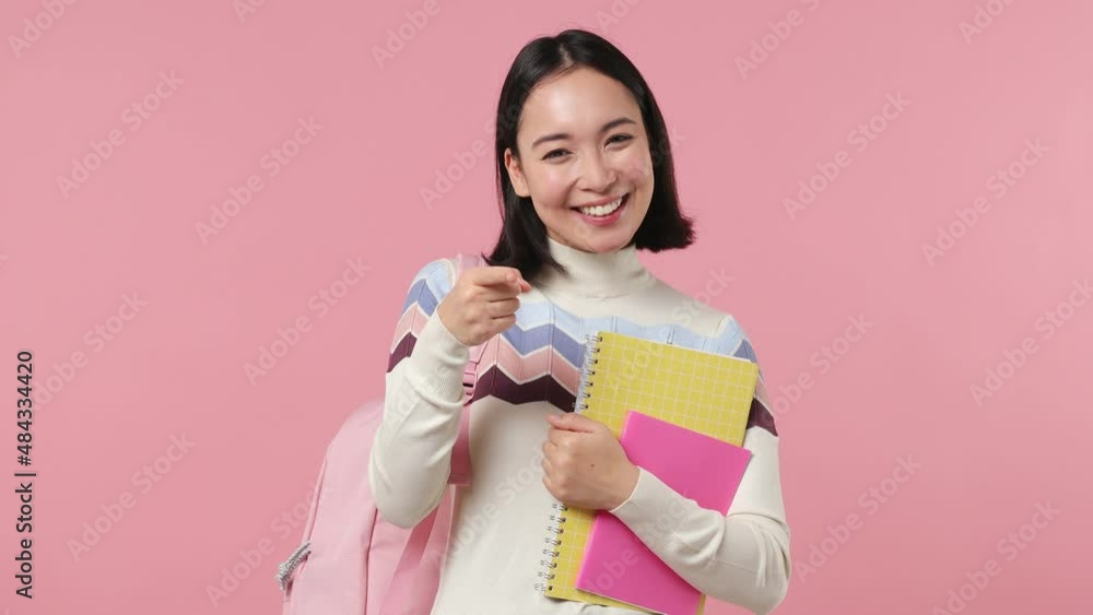 Girl teen student of Asian ethnicity wears shirt backpack hold books wink eye blink isolated on plain pastel light pink background studio portrait. Education in high school university college concept Girl teen student of Asian ethnicity wears shirt backpack hold books wink eye blink isolated on plain pastel light pink background studio portrait. Education in high school university college concept
