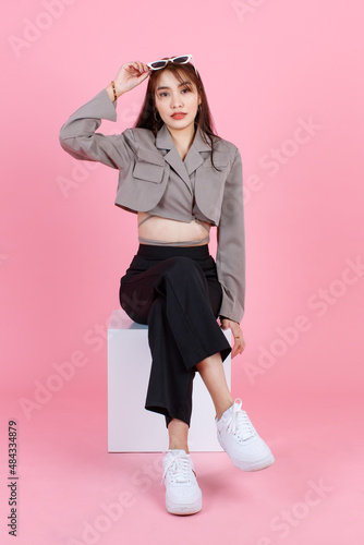 Portrait studio shot of Asian young urban trendy female hipster teenager fashion model in casual crop top street wears jacket holding sunglasses on head sit on white square box stool 