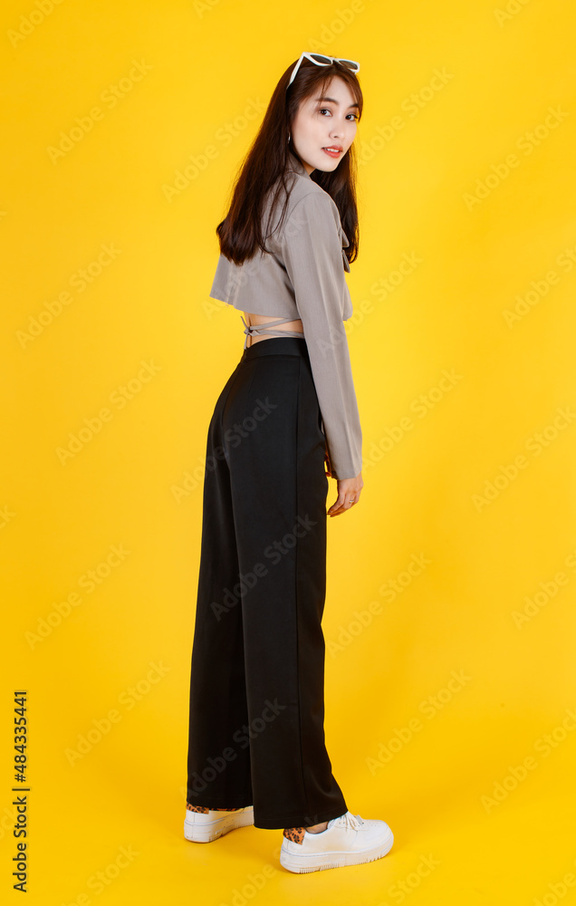 Fototapeta premium Portrait studio full body shot of Asian urban trendy modern fashionable long hair female hipster teen model in casual street wears crop top shirt standing posing look at camera on yellow background