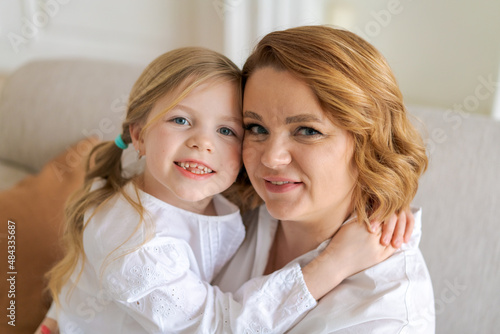 European mom and little daughter are hugging while sitting on sofa in bright living room at home. The relationship between children and parents. Happy motherhood