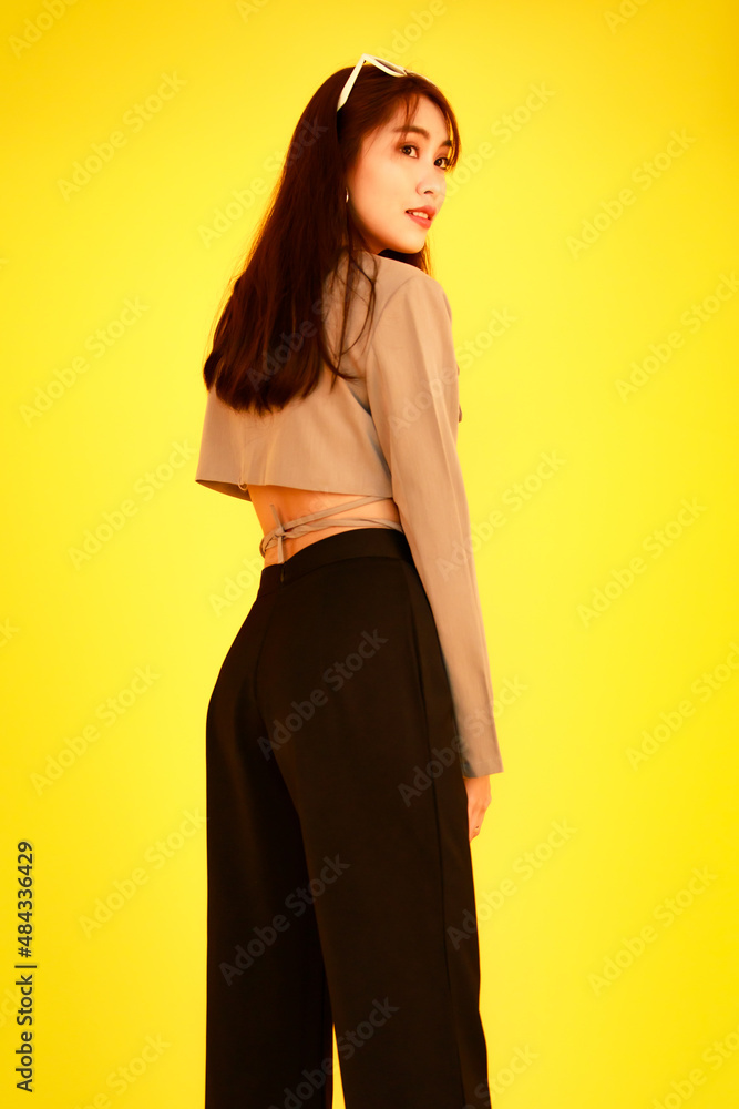 Naklejka premium Portrait studio shot of Asian trendy fashionable female hipster teen model in casual crop top street wears jacket sunglasses sneakers holding handbag purse look at camera on yellow background