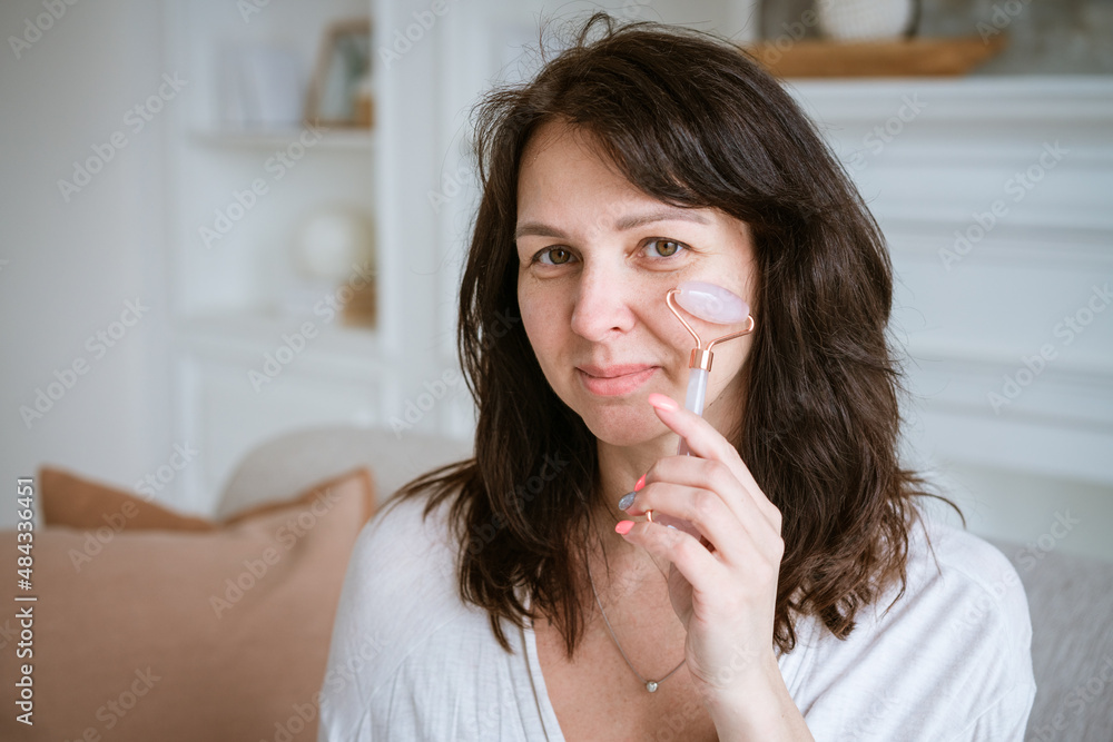 Brunette smiling woman 35 years old plus clean fresh face and hands with long hair makes facial massage with gouache roller in a bathrobe while sitting at home on the couch. Facial skin care concept Brunette smiling woman 35 years old plus clean fresh face and hands with long hair makes facial massage with gouache roller in a bathrobe while sitting at home on the couch. Facial skin care concept