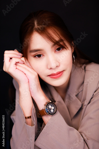 Portrait closeup studio shot of Asian young glamour trendy urban fashionable female model wearing makeup in casual fashion shirt wrist watch earring sitting smiling look at camera on black background