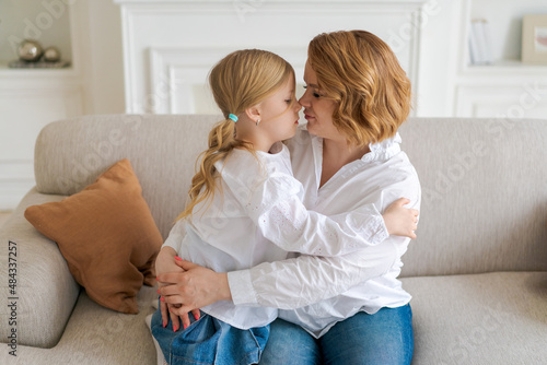 European mom and little daughter are hugging while sitting on sofa in bright living room at home. The relationship between children and parents. Happy motherhood