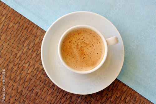 Cup of cappuccino coffee in a white saucer on wooden table with a tablecloth, top view
