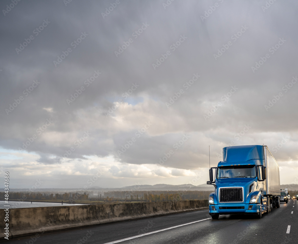 Blue big rig semi truck transporting cargo in dry van semi trailer running on the wet highway road with stormy rain sky