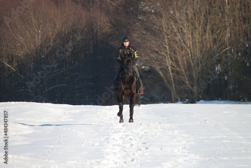 Ein Reiter mit einem schwarzen Pferd beim Ausritt im Schnee im Galopp an einem sonnigen Winter Tag frontal auf einen zukommend