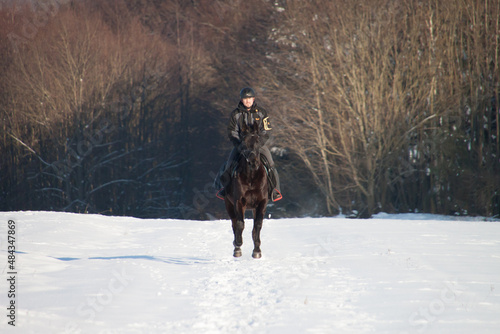 Reiter galoppiert auf schwarzem Pferd einen Weg im Schnee frontal in der Sonne vor einem Wald den Weg hoch und freut sich