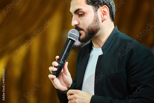 A charming young man leading with a microphone in a black suit.