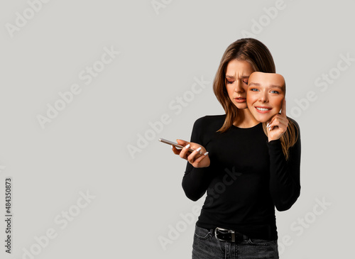 Portrait of a sad girl with a smartphone in one hand and a smiling mask in the other hand.