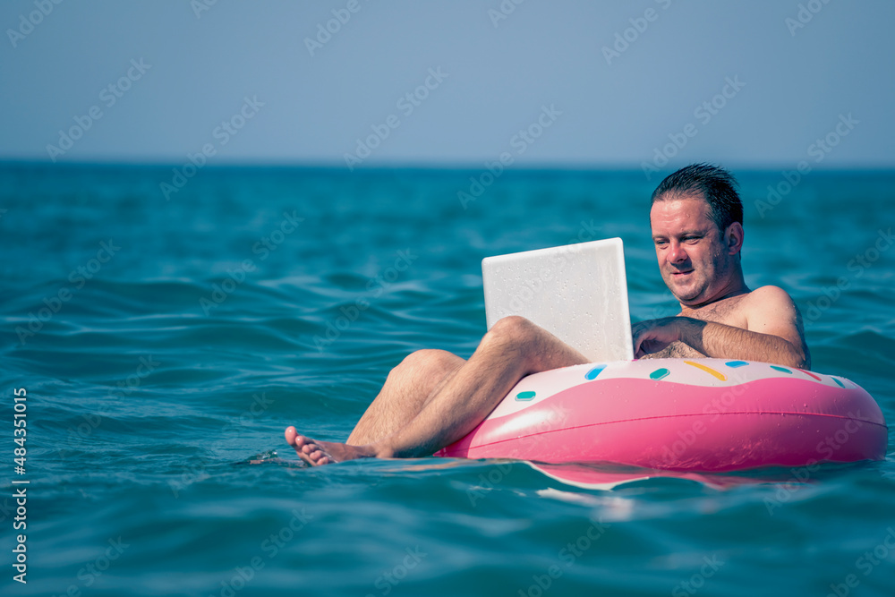 Working anywhere and any time concept. Young man working, using laptop computer on inflatable ring in the water. Freelance work, vacations, distance work, social distancing. Copy space.