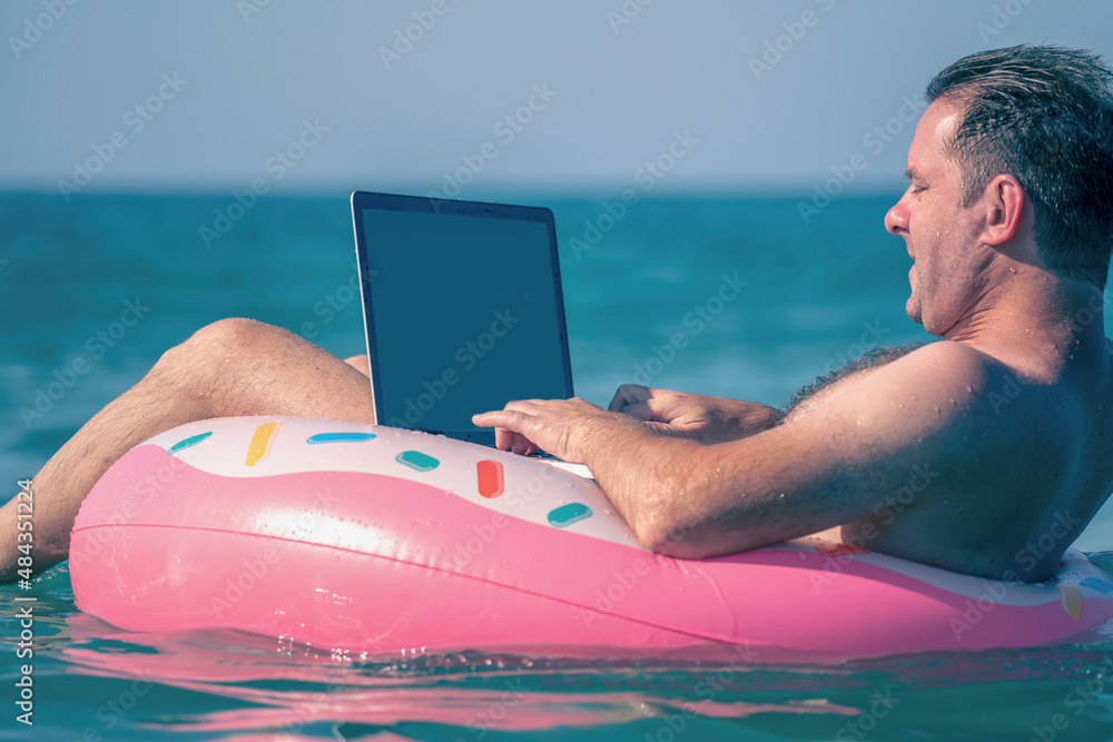 Young man working, using laptop computer on inflatable ring in the water. Freelance work, vacations, distance work, social distancing, e-learning, connection concept.